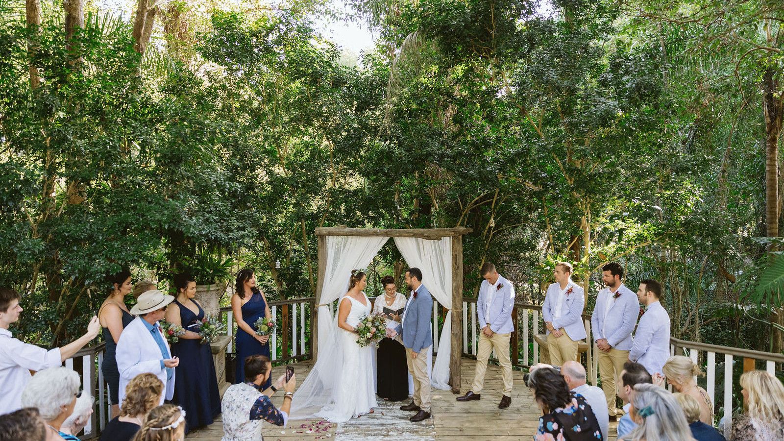 A wedding ceremony seen from behind the seated guests, Felicity with the couple under an arch of white voile on a timber deck beneath rainforest trees