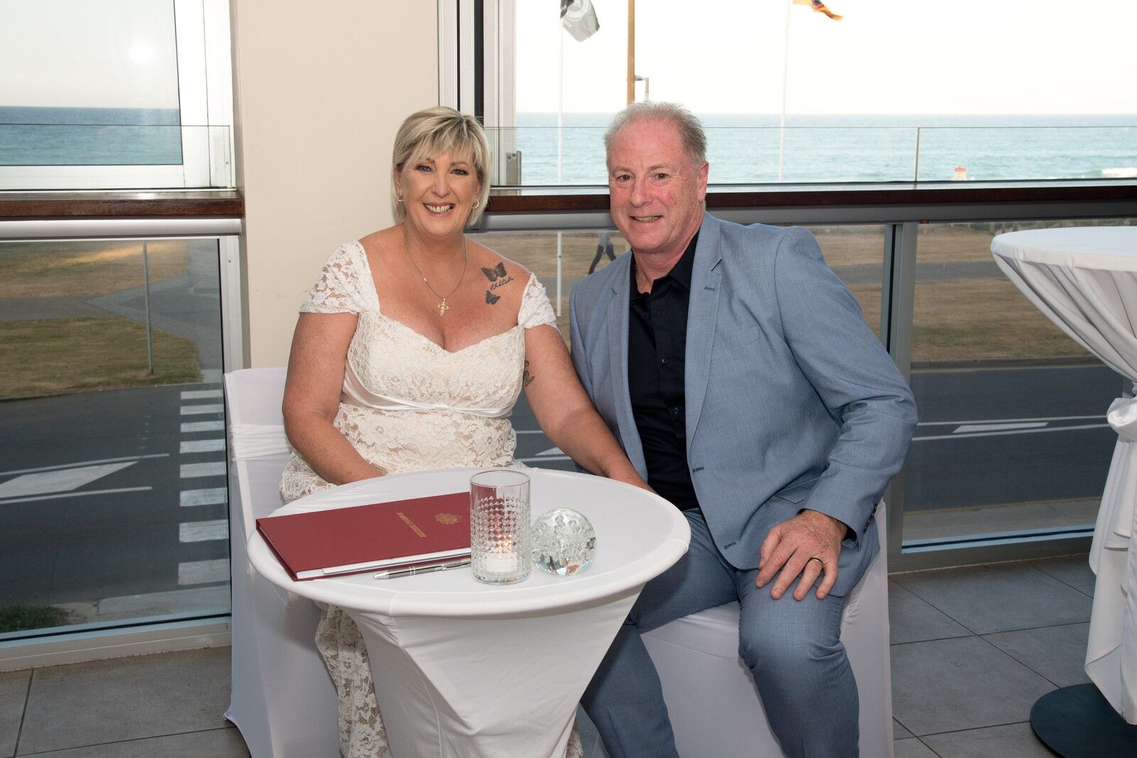 The couple seated at a white table with their marriage certificate in a red folder in front of them, the beach and the ocean through the window behind