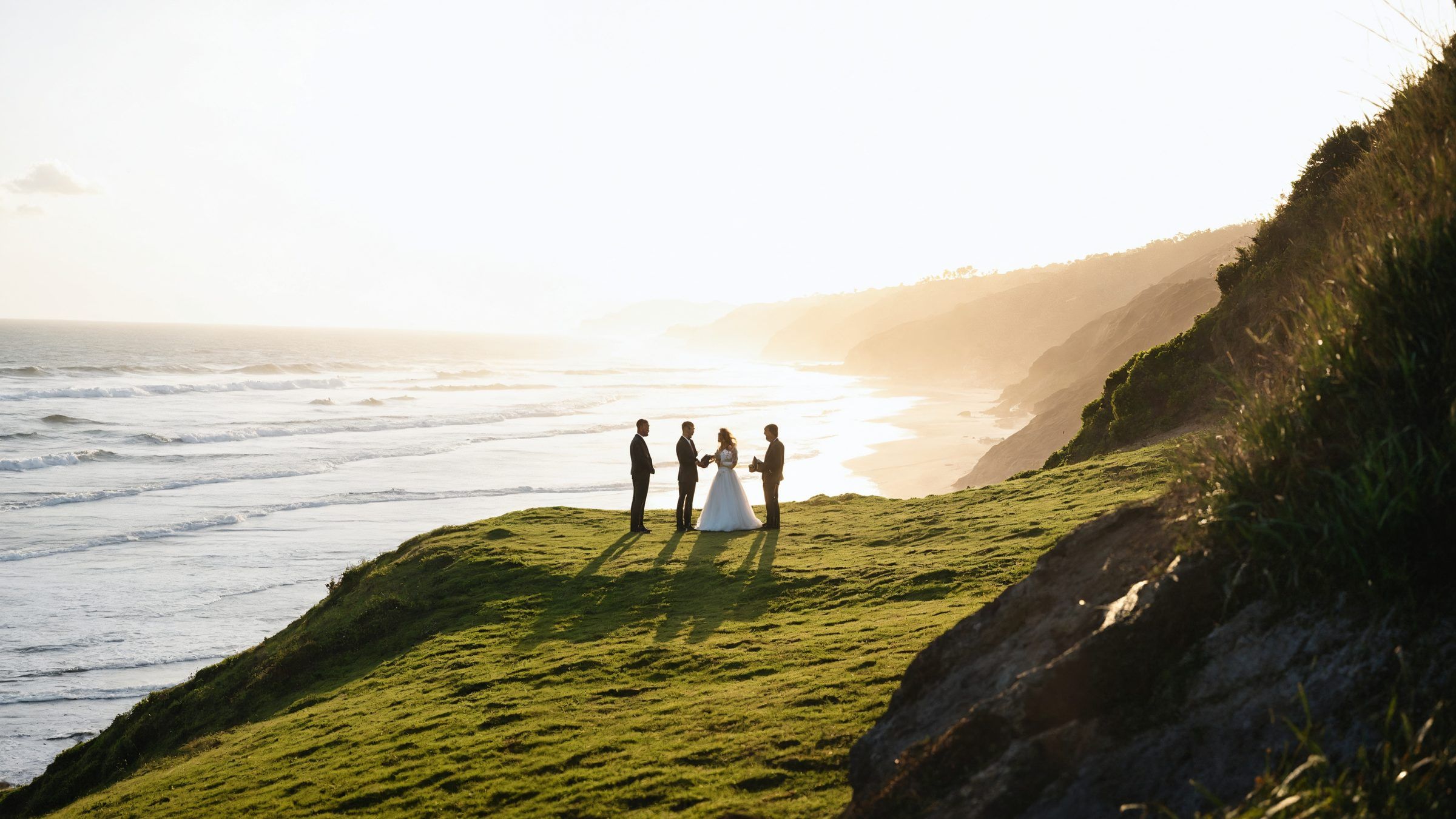 Four figures standing on a grassy headland at sunrise, tiny against a long surf beach and the sea