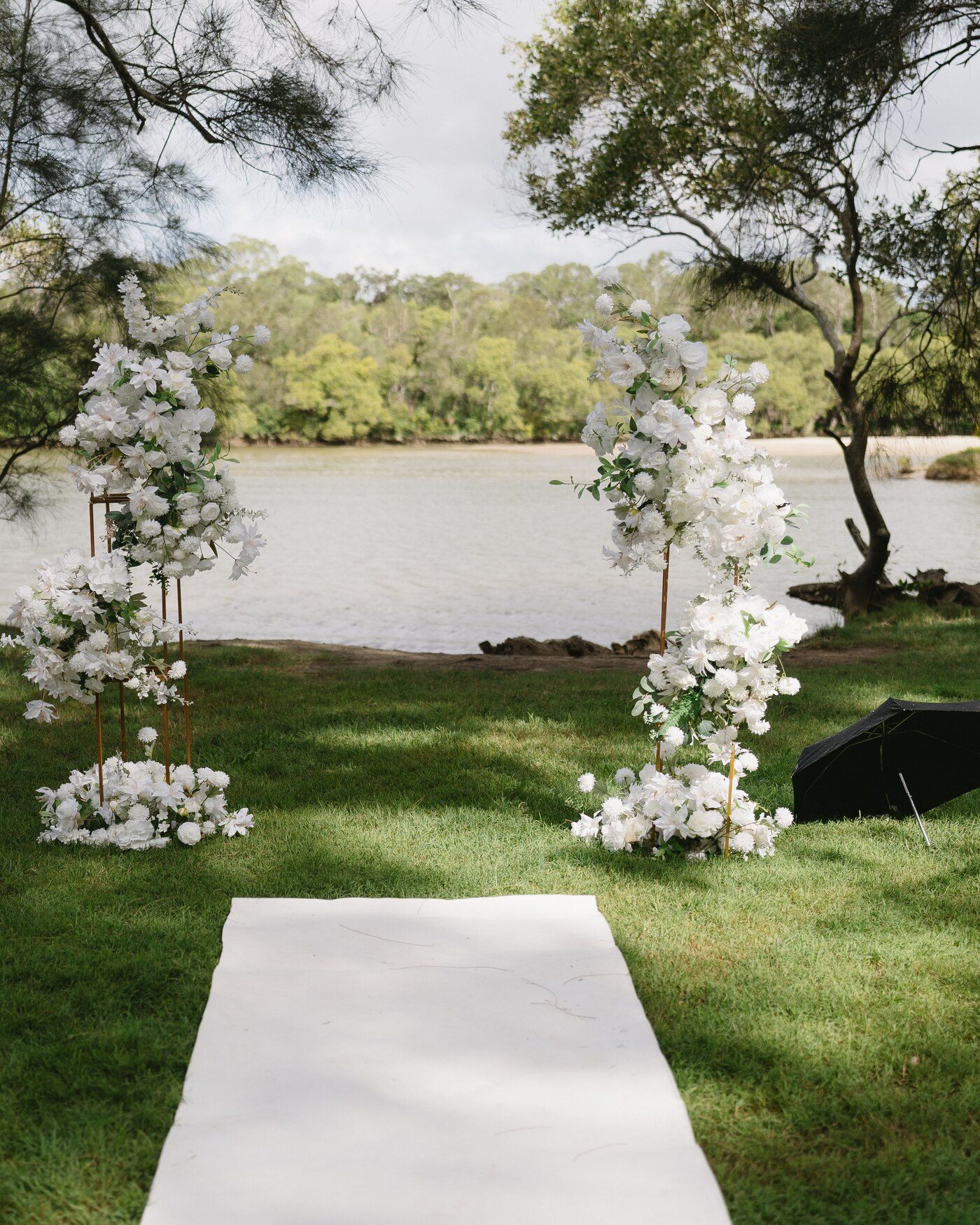 A white aisle runner on the lawn leading to a white flower arch at the edge of a creek, casuarinas overhead