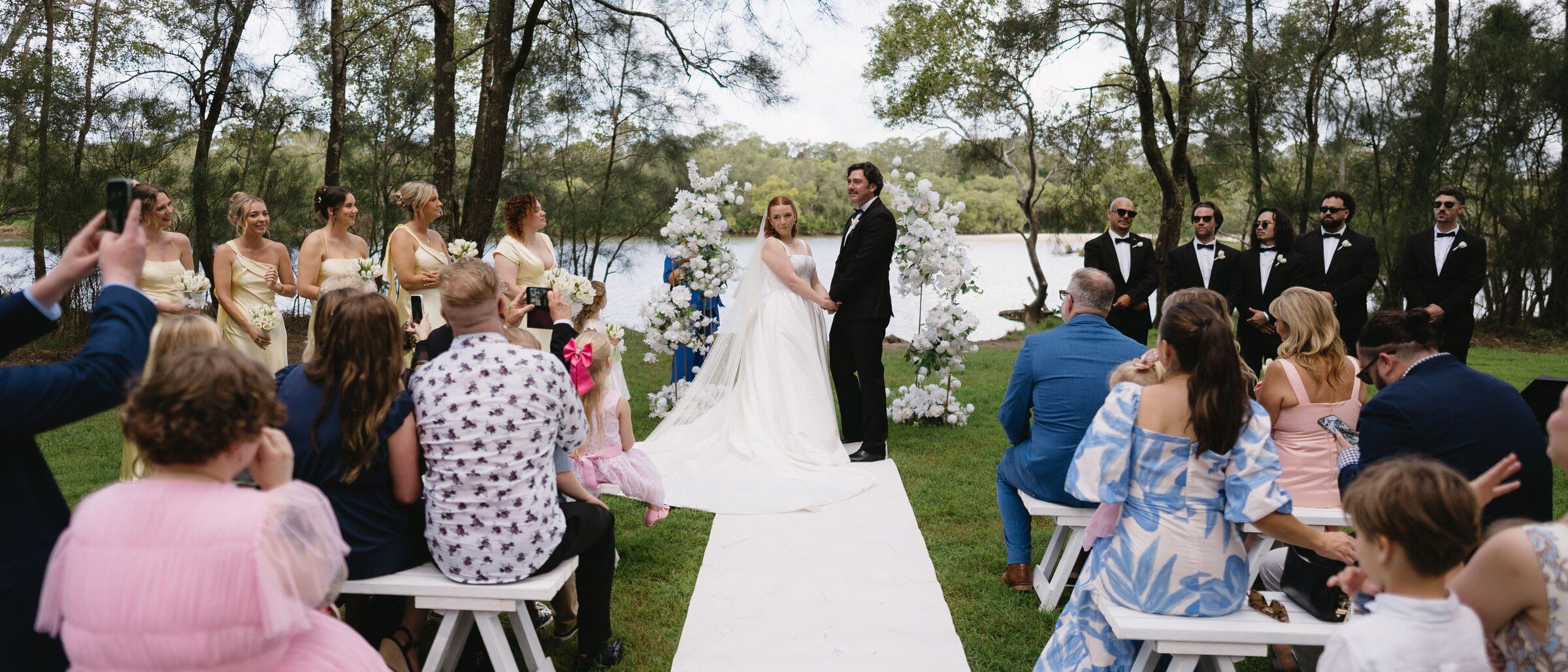 A ceremony on a creek bank seen from behind the seated guests, the couple and Felicity under a white flower arch and the wedding party either side