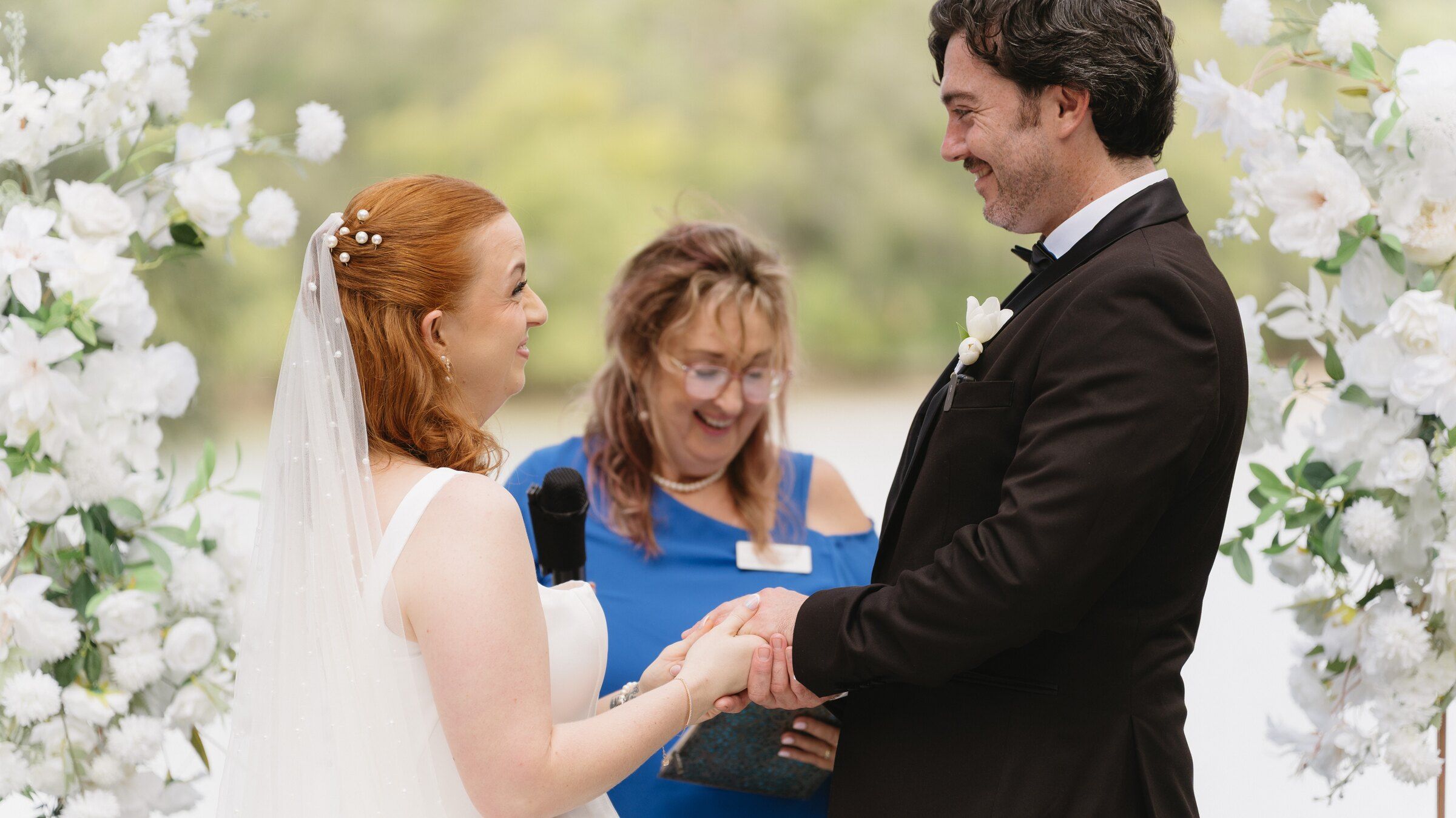 The bride and groom holding hands under a white flower arch, Felicity laughing between them with her microphone and book, the creek behind