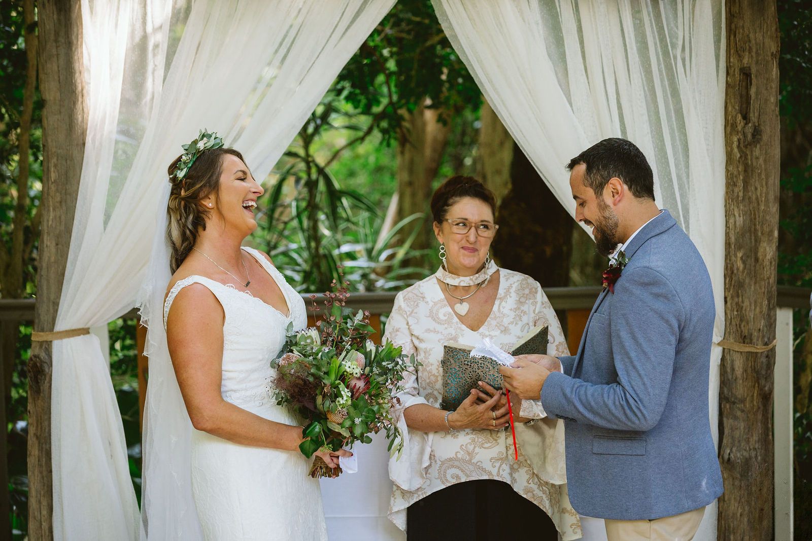 Felicity between the bride and groom under a timber arch hung with white voile, the bride laughing, Felicity reading from her folder