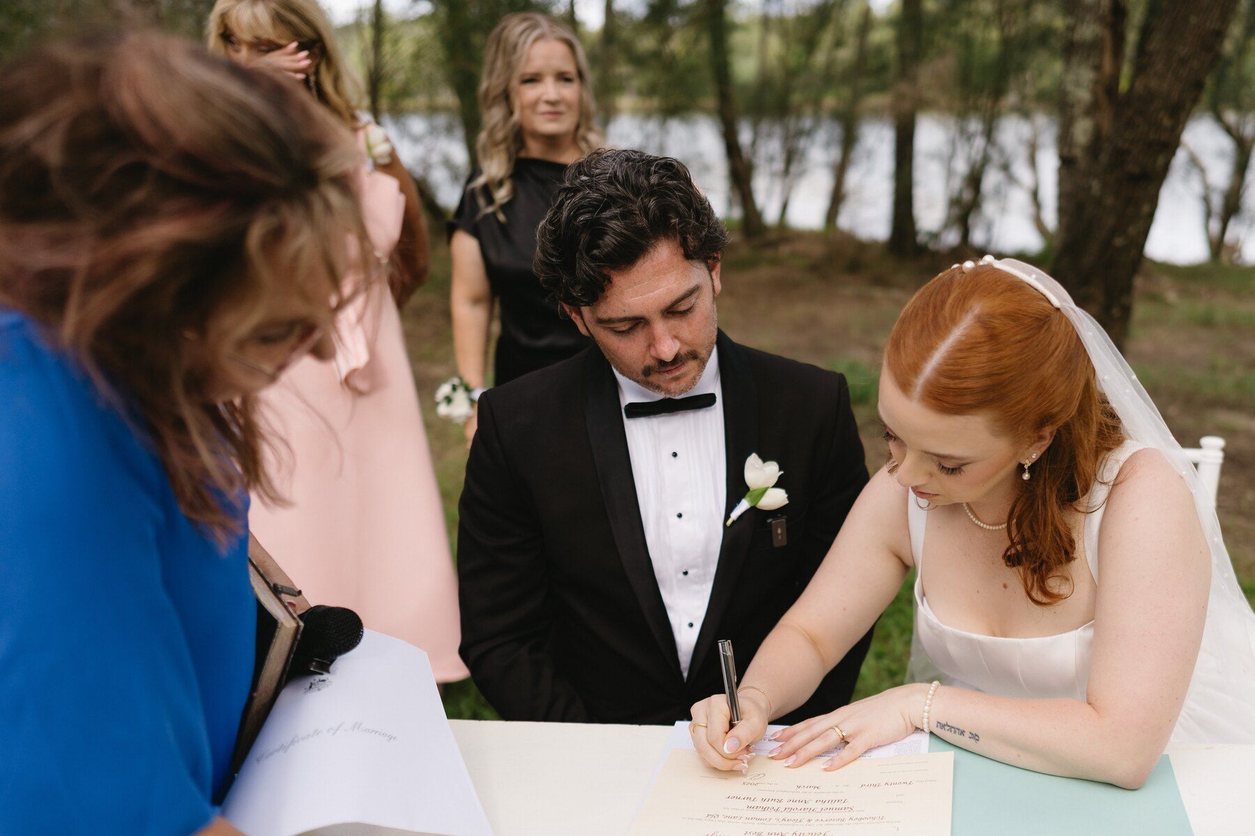 The bride signing the marriage certificate at a white table while the groom watches, Felicity in the foreground holding the paperwork, the creek through the trees behind