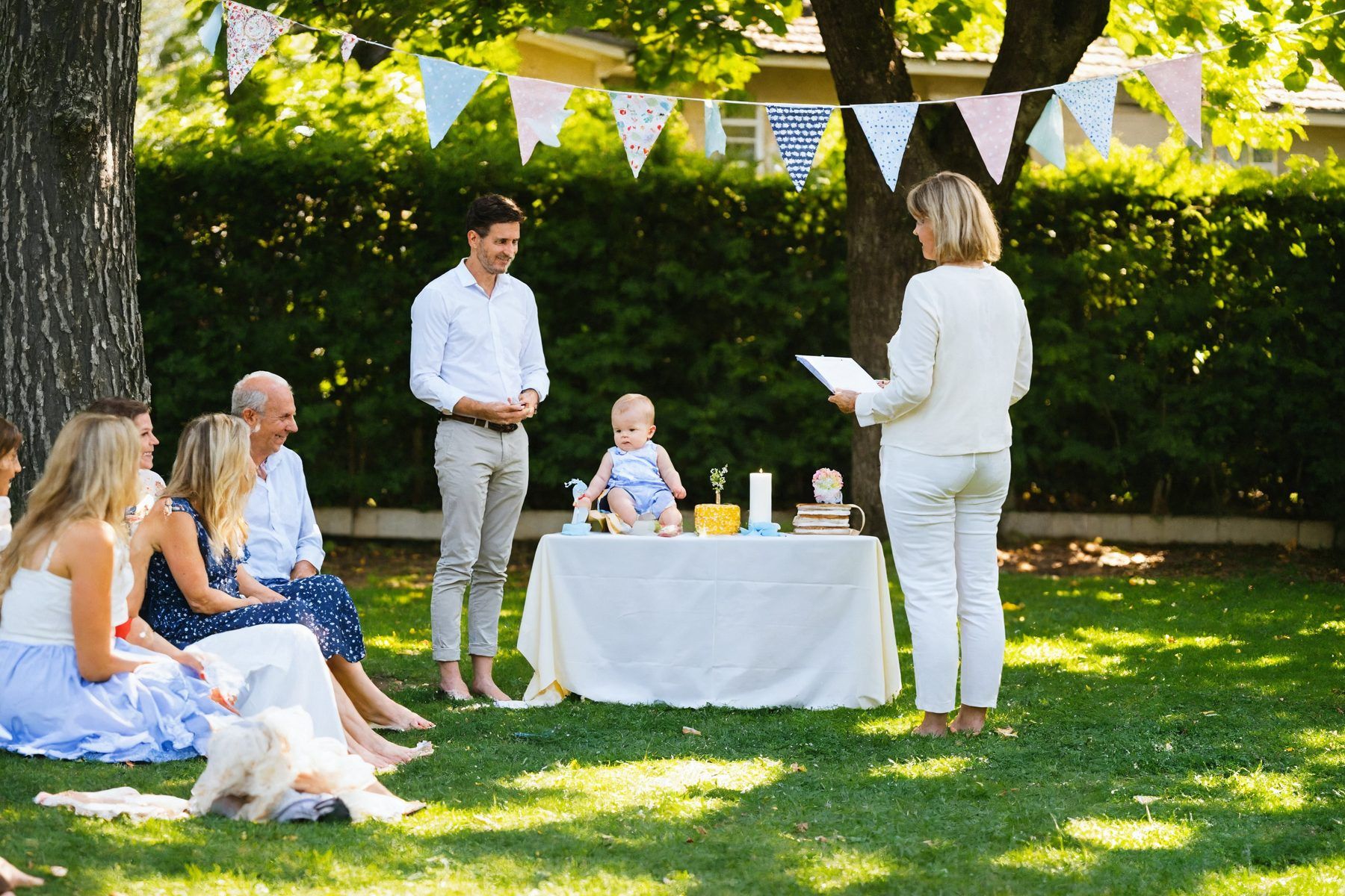 A naming ceremony in a backyard, bunting strung between the trees, the baby on a cloth-covered table and the family gathered round