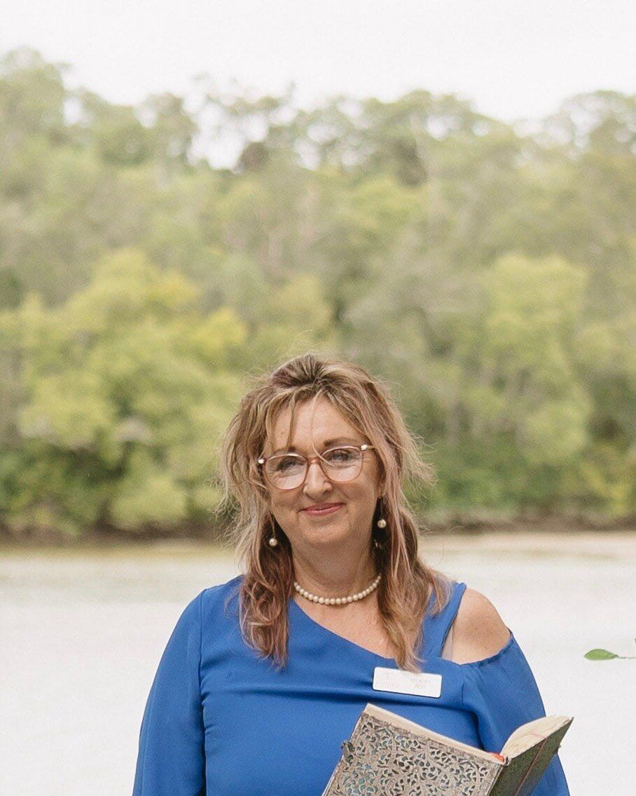 Felicity smiling beside a river, in a blue off-the-shoulder top and pearls, her name badge on and an open ceremony book in her hands