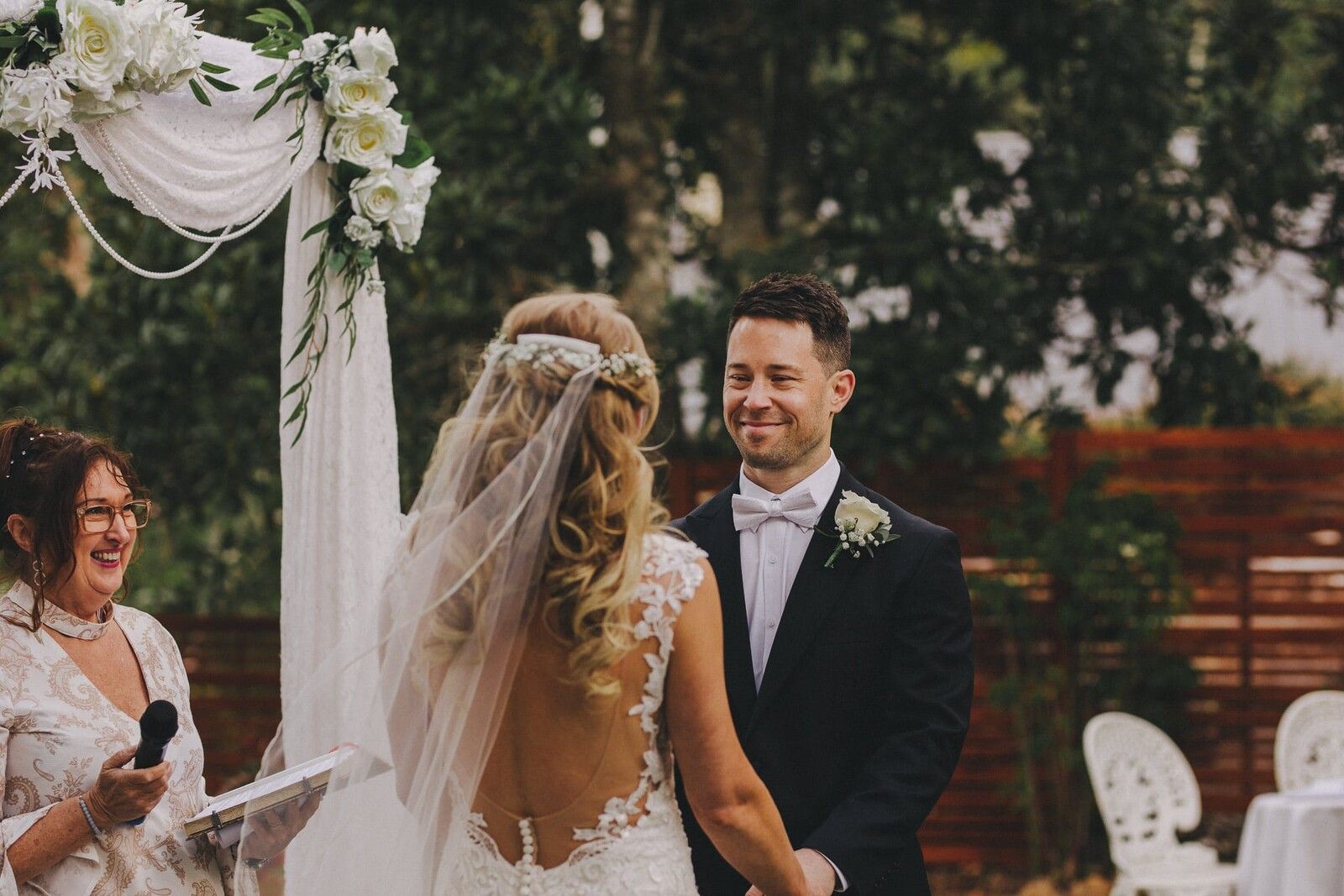 A bride and groom facing each other under a floral arch as the vows are spoken, the groom smiling, the celebrant at the edge of the frame with a microphone