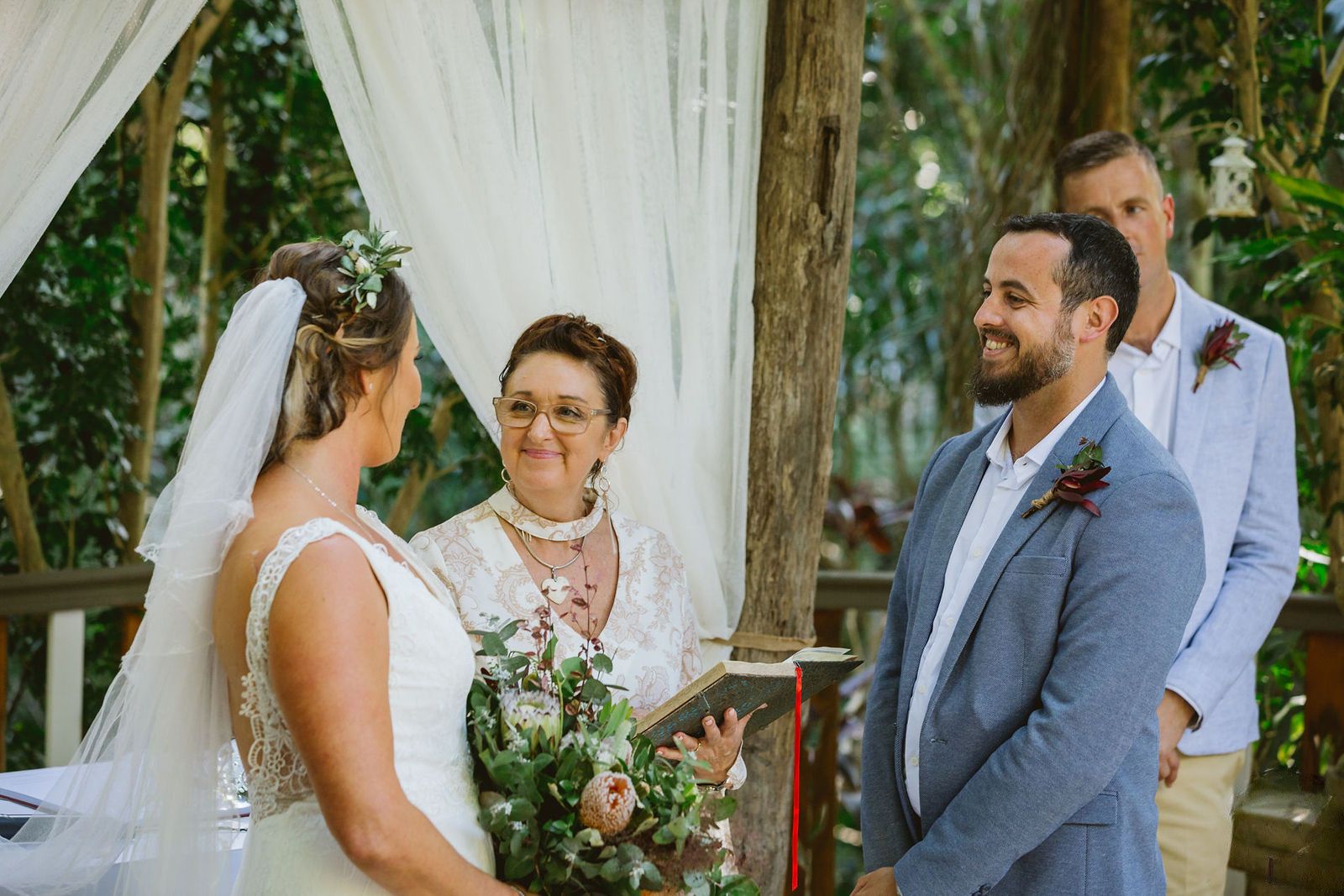 Felicity smiling as she reads the ceremony from her folder, the bride's veil and native bouquet on one side, the groom on the other