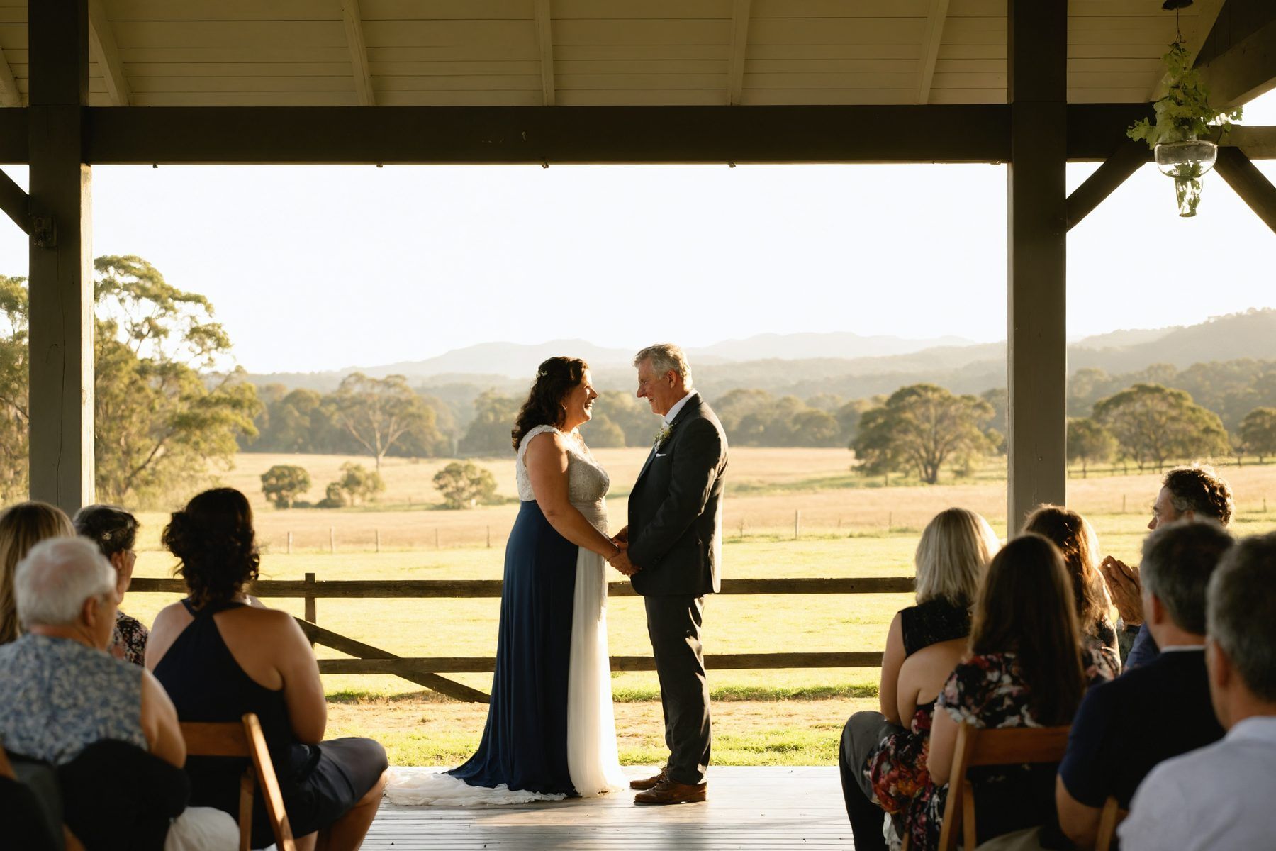 A couple in their fifties renewing their vows on a farmhouse verandah, family seated either side and paddocks behind