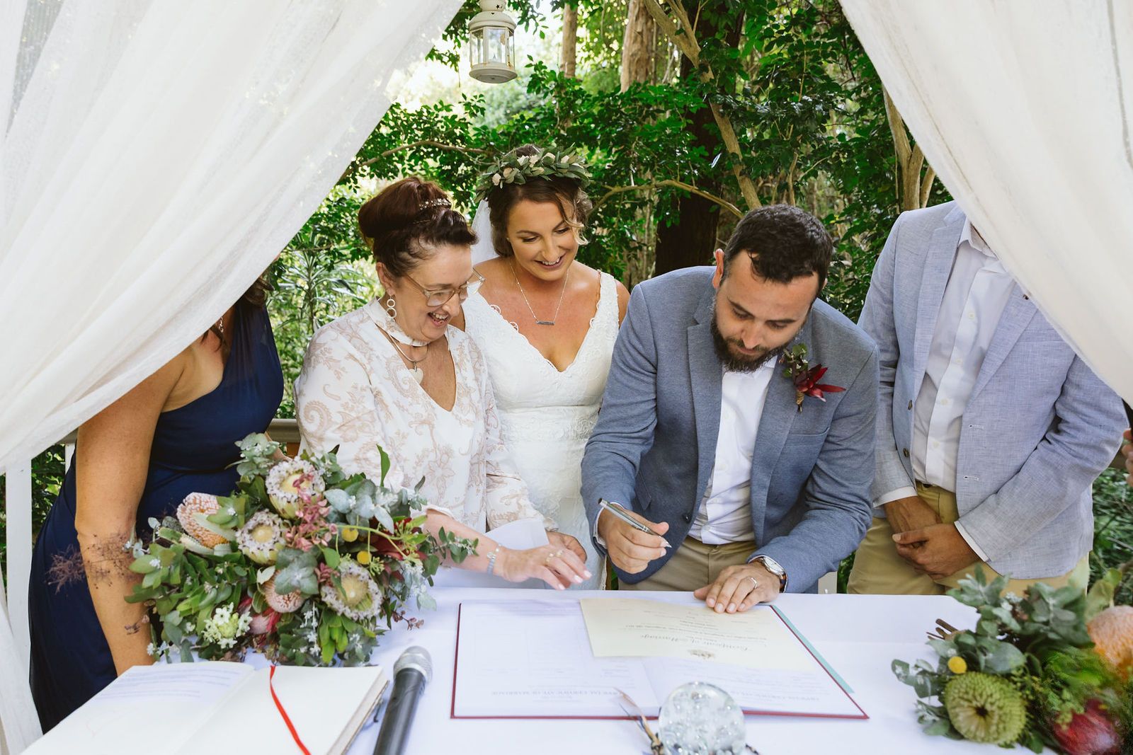 Felicity at the signing table as the groom signs the register, the bride and witnesses leaning in, native flowers on the white cloth