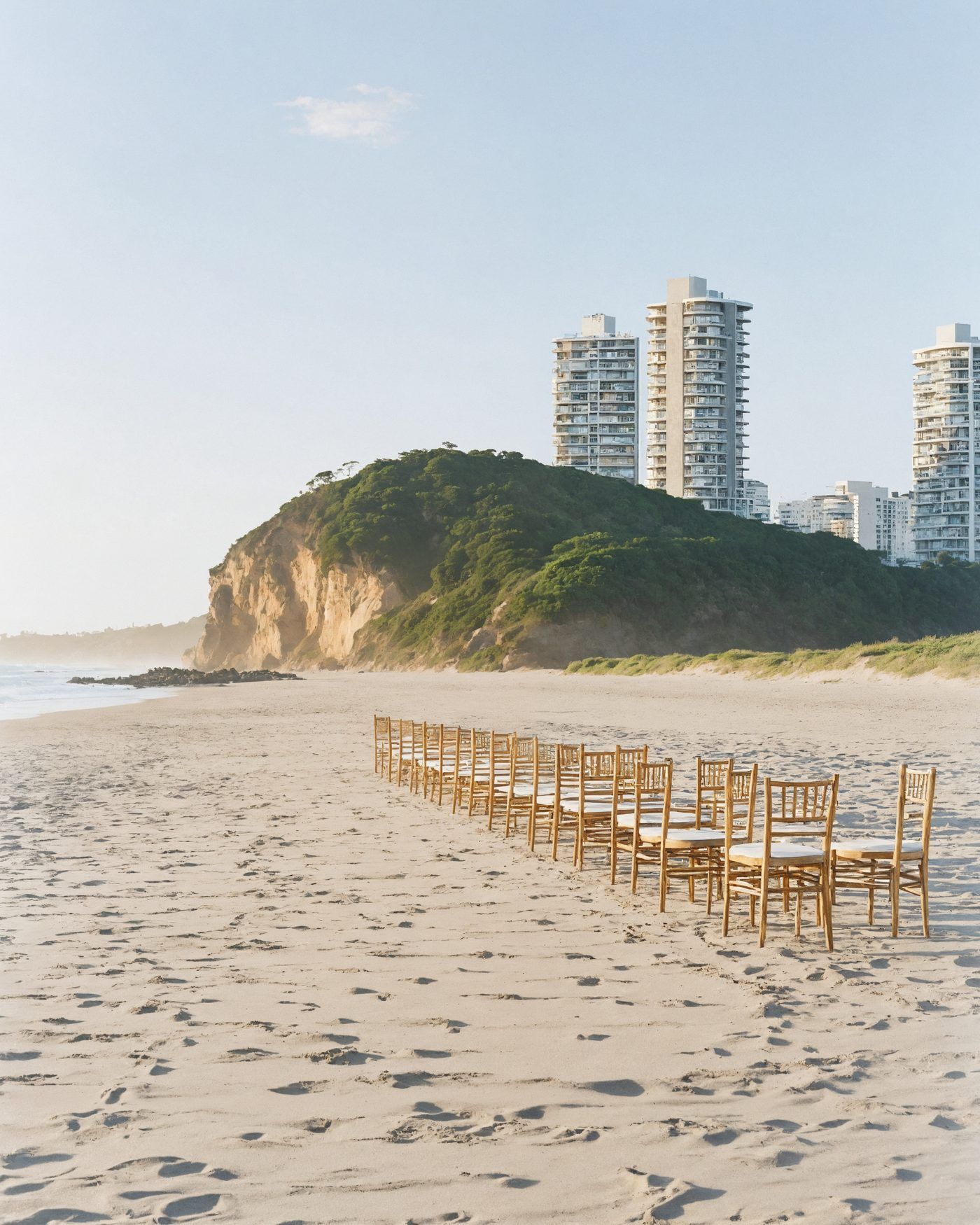 Timber chairs set on the sand at a border beach, a green headland and the distant Gold Coast towers behind in morning light