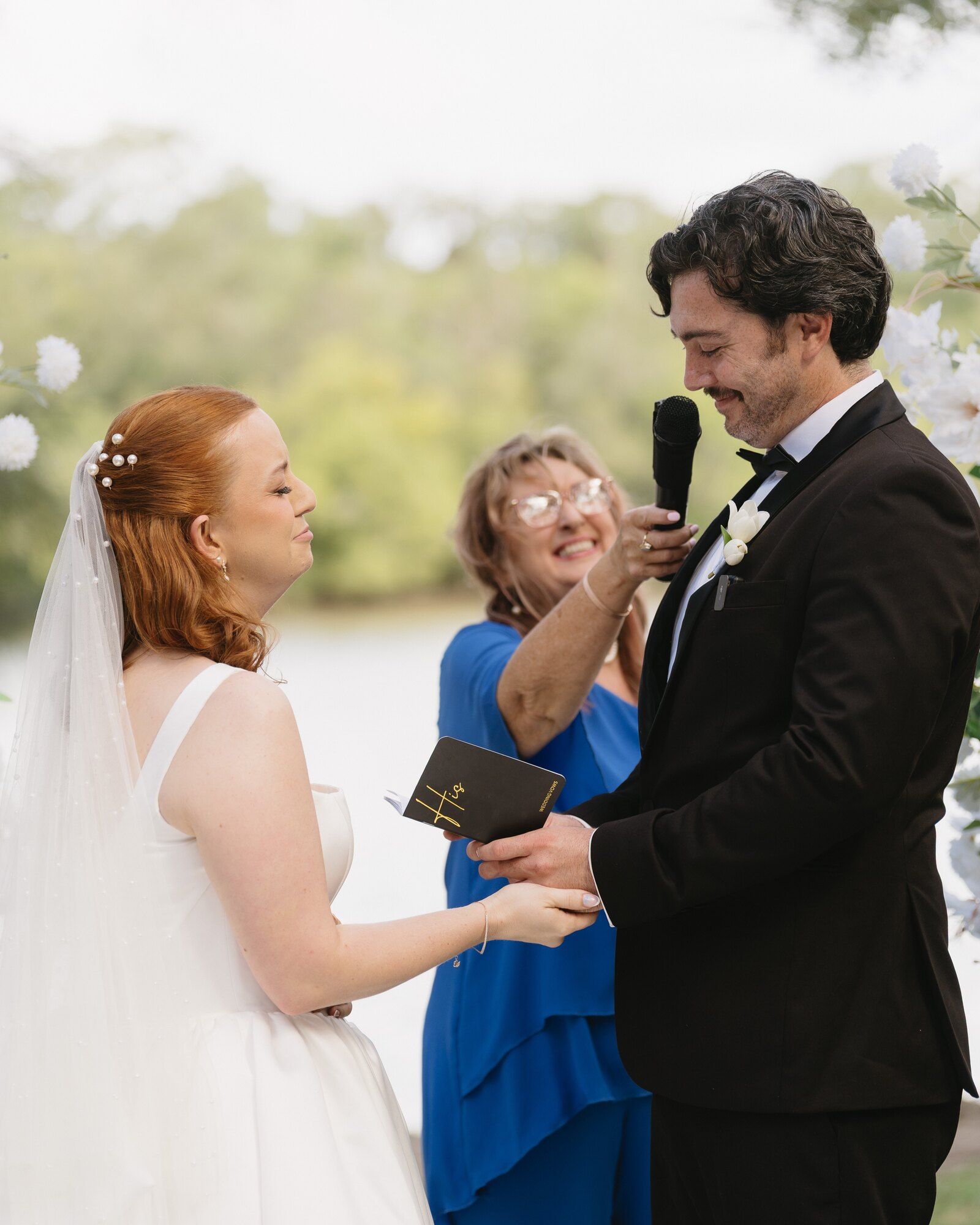 Felicity holding a microphone up for the groom as he reads his vows from a small black book, the bride smiling beside him and the creek behind